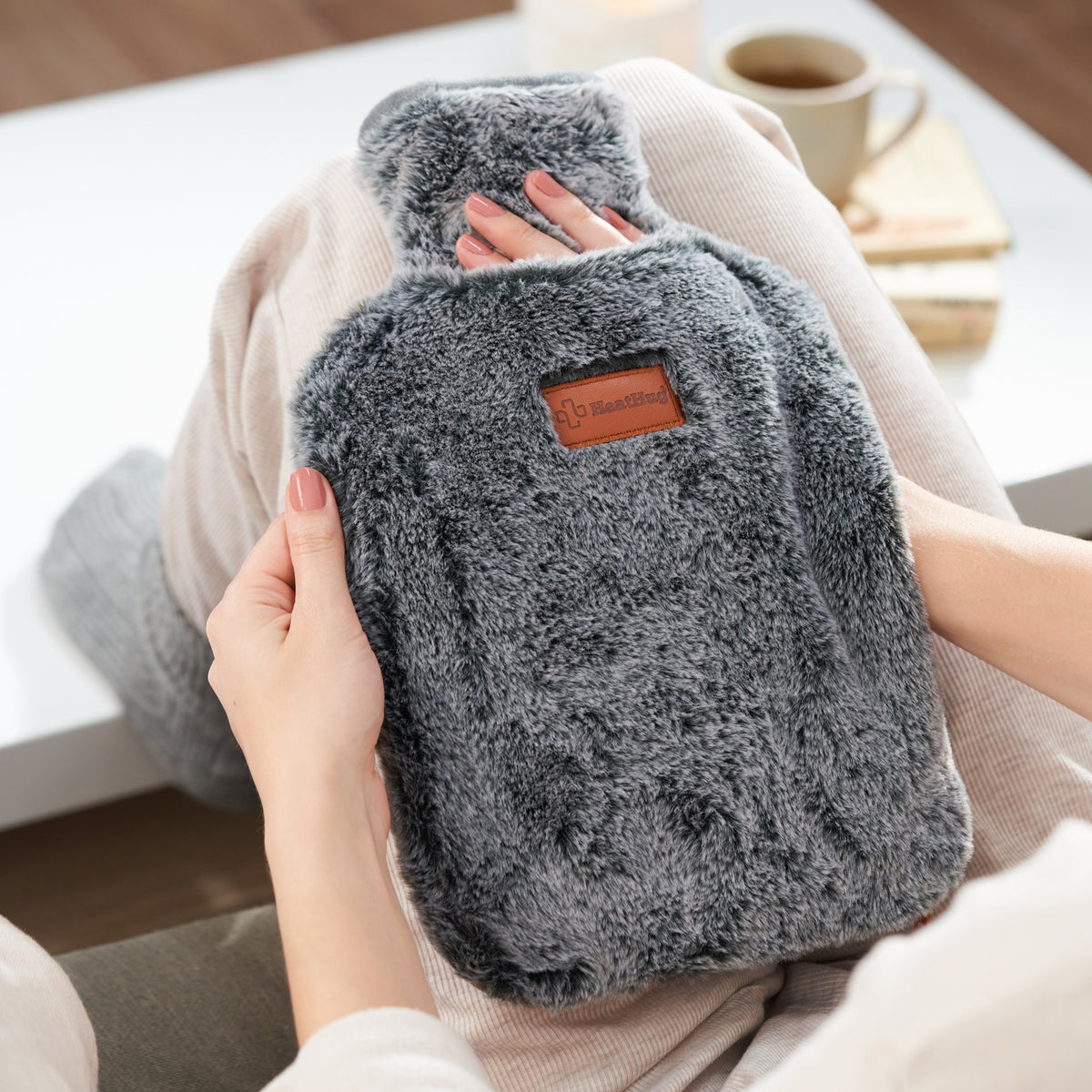 BigHug 2l hot water bottle with grey faux fur cover, held on a lap by a coffee table